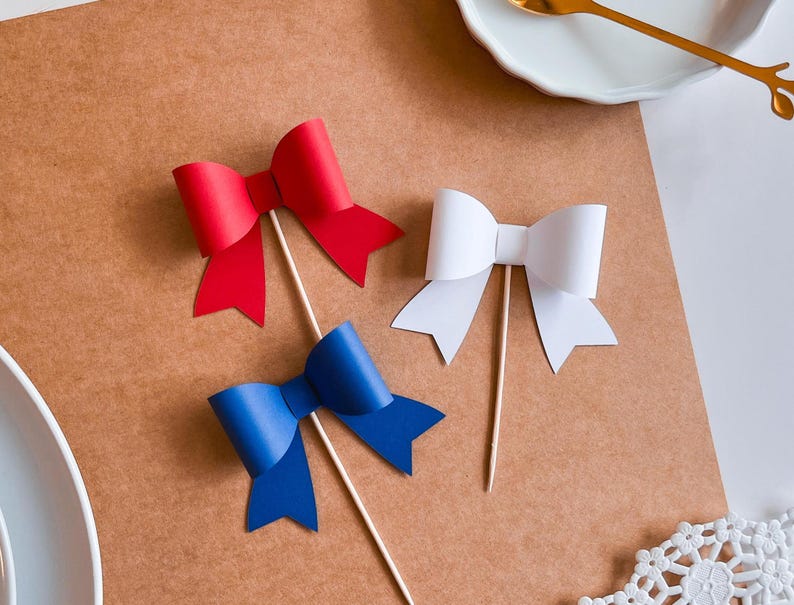 May include: Three paper bows on sticks, one red, one white, and one blue. The bows are arranged on a brown surface.
