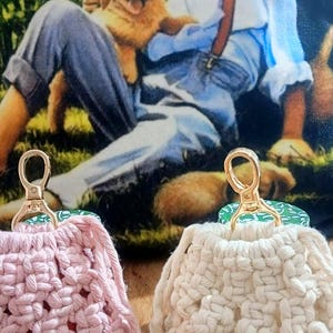 May include: Two small macrame pouches with gold-toned lobster clasps. One pouch is light pink and the other is off-white. Both feature a knotted cord design, drawstring closures with wooden beads at the ends, and a textured, woven appearance.
