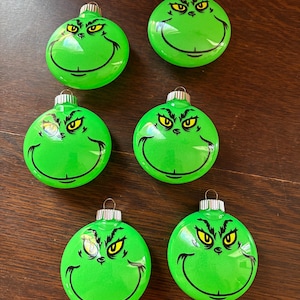 Peut inclure: Six boules de Noël rondes et vert vif, chacune arborant le motif imprimé d'un visage avec des yeux jaunes, des sourcils noirs et un large sourire malicieux. Chaque boule possède un capuchon en métal argenté sur le dessus pour la suspendre.