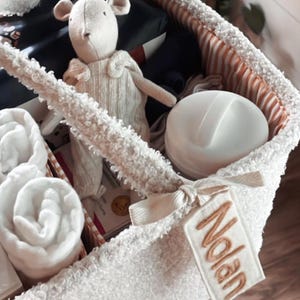 Puede incluir: Una cesta blanca texturizada llena de artículos para bebés. Incluye un peluche, toallas enrolladas y un recipiente blanco. Una etiqueta bordada con el nombre "Nolan" está adjunta a la cesta.