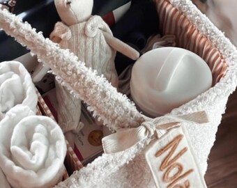 May include: A white, textured basket filled with baby items. Includes a stuffed animal, rolled towels, and a white container. A tag embroidered with the name "Nolan" is attached to the basket.