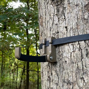 May include: A tan-colored tree hook with a black rubberized grip, secured to a tree trunk with a black nylon strap. The hook features a hinged design with a visible metal bolt and a small engraved logo on the side of the mounting bracket.