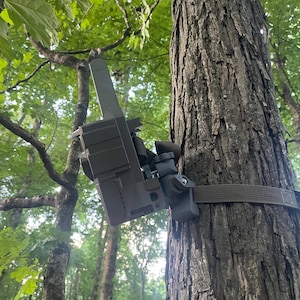 May include: A trail camera is mounted on a tree trunk with a brown strap. The camera is a muted green color and is attached to the tree in a forest setting. The background includes green leaves and other trees.