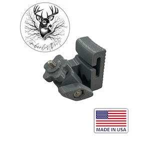 May include: A gray plastic bracket with a screw and bolt, likely for mounting. The bracket has a unique shape with a slot and a textured surface. The image also includes a circular logo with a deer and the text "Timberlost Pro" and a "Made in USA" sticker.