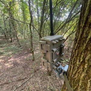 May include: A tan trail camera with a black antenna mounted to a tree trunk using a gray adjustable bracket and a strap. The rectangular device features a rugged, hinged casing with locking latches on the side and a central label on the back.