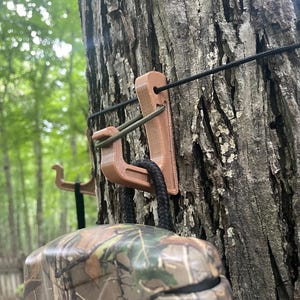 May include: A light brown, 3D-printed plastic hook mounted to a tree trunk. The hook features a vertical slot for a black cord and a curved arm supporting a camouflage-patterned object hanging by a black braided strap from the hook's base.