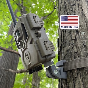 May include: A matte olive green trail camera with a black antenna, secured to a tree trunk by a gray adjustable mount and a brown strap. A rectangular sticker with the American flag and the text MADE IN USA is visible on the tree bark.