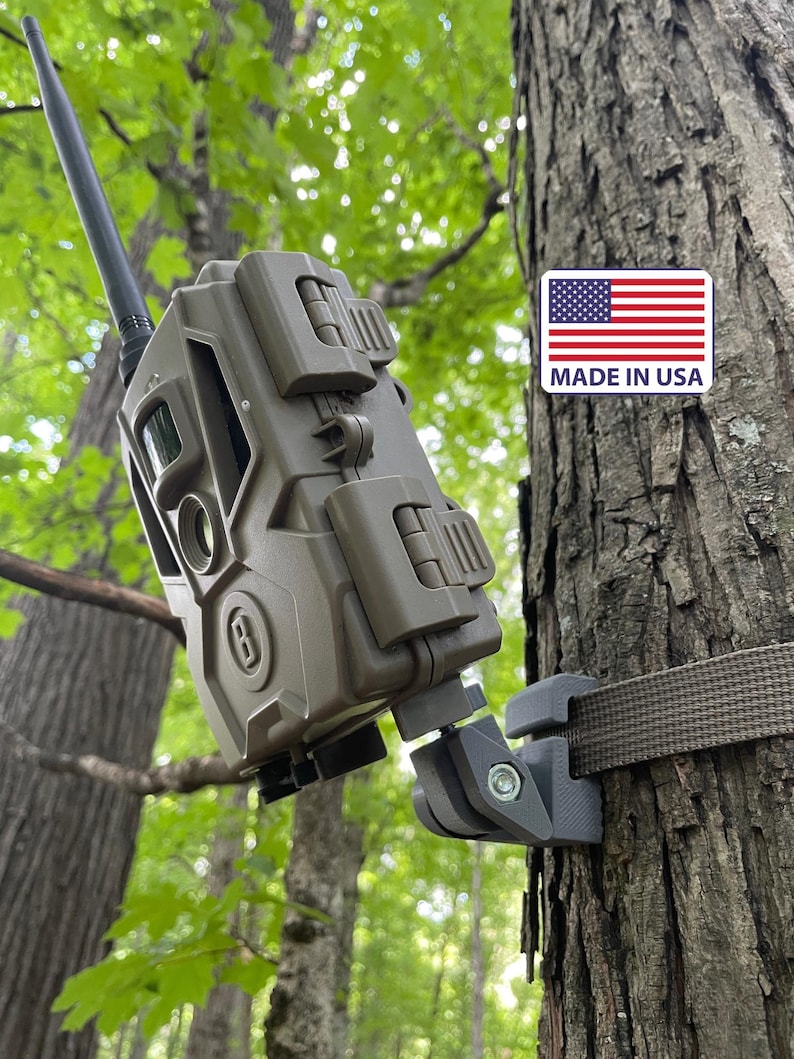 May include: A matte olive green trail camera with a black antenna, secured to a tree trunk by a gray adjustable mount and a brown strap. A rectangular sticker with the American flag and the text MADE IN USA is visible on the tree bark.