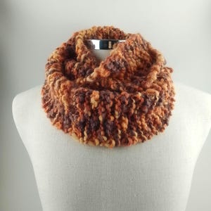 May include: A chunky knit infinity scarf in shades of orange, brown, and cream. The scarf is displayed on a mannequin, showcasing its textured, warm appearance. This cozy accessory is perfect for cold weather.