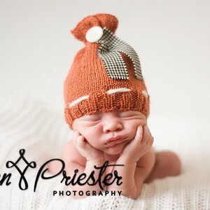 May include: A rust-coloured knitted beanie with a ribbed brim, a white horizontal stripe, and a top knot secured by a white button and a black and white houndstooth patterned ribbon. The text Jen Priester Photography is visible.