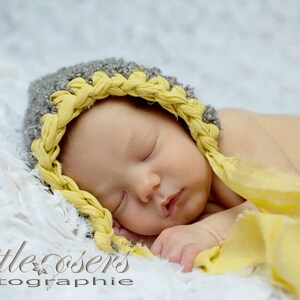 May include: A sleeping baby wearing a grey and yellow crocheted hat. The baby is lying on a white fluffy blanket.