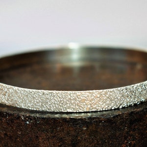 May include: A silver bangle bracelet with a textured surface. The bracelet is sitting on a brown surface.