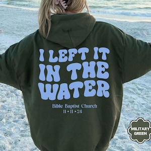 May include: Military green hoodie with the text "I LEFT IT IN THE WATER" in light blue, along with "Bible Baptist Church 11-11-24". The hoodie is a relaxed fit, perfect for casual wear.