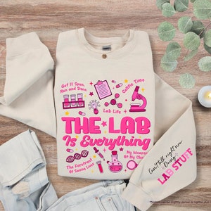 May include: A beige sweatshirt with a pink graphic design that says "The Lab is Everything" and includes various science-related icons. The text "Get It Spun, Run and Done" is also included in the design.