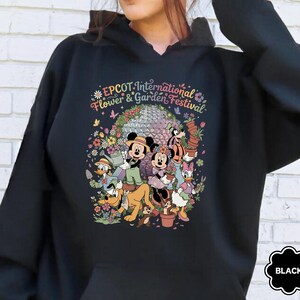 May include: Black hooded sweatshirt featuring a colorful graphic of Disney characters at the EPCOT International Flower & Garden Festival. The design includes Mickey and Minnie Mouse, Donald Duck, and Pluto, surrounded by flowers. The word "BLACK" is in a black bubble.