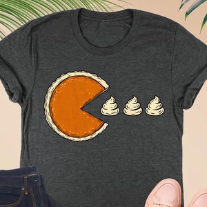 May include: Dark grey heather t-shirt featuring a pie slice design with three dollops of whipped cream. The shirt is paired with dark blue jeans, a pink wallet, and pink sneakers. The shirt's design is a playful take on a classic video game.