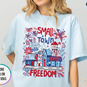 May include: Light blue t-shirt with a colorful graphic featuring the words "Small Town Freedom" and a town scene with buildings, flowers, and a water tower. The shirt has short sleeves and a round neckline.