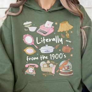 May include: A military green hooded sweatshirt featuring a graphic print with the text "Literally from the 1900's" surrounded by illustrations of a pink typewriter, a lamp, a mirror, a casserole dish, a kettle, a rotary phone, and a shopping cart.