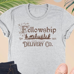Puede incluir: Una camiseta gris jaspeada con un gr&aacute;fico marr&oacute;n de los personajes de la Comunidad del Anillo caminando en fila. El texto "Fellowship" est&aacute; por encima de los personajes y "EST 1954 fast, reliable, precious DELIVERY CO." est&aacute; por debajo de los personajes.