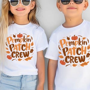 May include: Two white short-sleeved t-shirts featuring a graphic print that reads Pumpkin Patch Crew in orange and brown lettering, surrounded by illustrations of small pumpkins and autumn leaves in shades of orange, brown, and yellow.