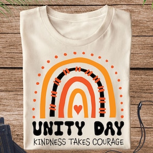 May include: Cream-colored t-shirt featuring a rainbow design in orange and black, with a heart at the center. The text "UNITY DAY KINDNESS TAKES COURAGE" is printed below the rainbow. The shirt is folded and placed on a wooden surface.