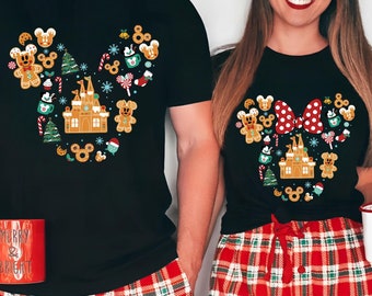 May include: A couple wearing matching black t-shirts with a gingerbread Mickey Mouse design. The shirts feature a gingerbread castle, candy canes, and other Christmas-themed treats. The couple is wearing red and green plaid pajama pants.