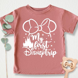 Pu&ograve; includere: Una maglietta color malva con un design grafico bianco che dice "My first Disneptrip" con una silhouette bianca del castello di Cenerentola e un fiocco bianco con orecchie da topo.