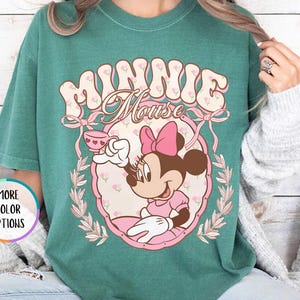 May include: A light green t-shirt with a vintage-style graphic of Minnie Mouse holding a teacup. The graphic features the text "Minnie Mouse" in a retro font.