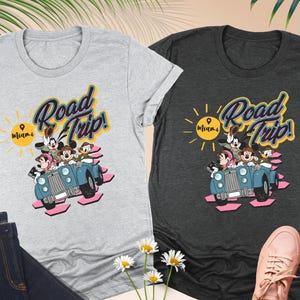Op de afbeelding: Twee grijze t-shirts met een "Road Trip! Miami"-afbeelding met stripfiguren in een blauwe auto. De shirts worden gecombineerd met een donkerblauwe jeans en roze sneakers. De afbeelding bevat ook witte madeliefjes en palmbladeren.