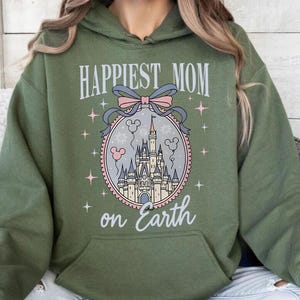 May include: Military green hoodie with a front graphic that reads "Happiest Mom on Earth." The graphic features a castle, balloons, and stars within a circular frame, with a bow at the top. The hoodie has a kangaroo pocket and a drawstring hood.