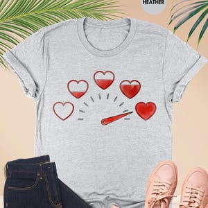 May include: A light gray t-shirt with a heart-themed graphic, paired with dark blue jeans, pink sneakers, and a small pink wallet. The shirt features a speedometer design with heart icons.