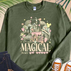 Pode incluir: Camisola verde militar com um gr&aacute;fico colorido que diz "The Most Magical Place". O design inclui um castelo, Mickey Mouse e outros elementos do parque tem&aacute;tico. Combinada com jeans azul escuro, t&ecirc;nis laranja e &oacute;culos.