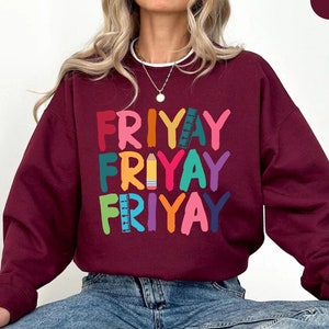 May include: A maroon sweatshirt with the word "FRIYAY" repeated three times in colorful letters with school supply graphics. The sweatshirt is being worn by a person sitting down.