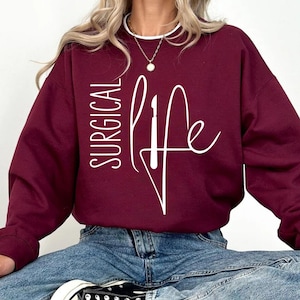 May include: A maroon sweatshirt with the text "SURGICAL LIFE" in white lettering. The text is stylized with a scalpel graphic.