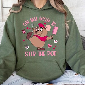 May include: Military green hoodie with a cartoon mouse graphic. The mouse is wearing a red shirt and hat, holding a spoon and a pink popsicle. The text "ON MY WAY TO STIR THE POT" is in pink.