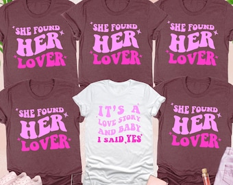 Op de afbeelding: Een verzameling t-shirts in heather maroon en wit. De maroon shirts tonen de tekst "SHE FOUND HER LOVER" in roze. Het witte shirt leest "IT'S A LOVE STORY AND BABY I SAID YES". Accessoires zijn schoenen, een wijnglas en een fles.