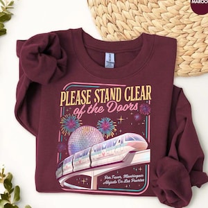 May include: Maroon sweatshirt with the text "PLEASE STAND CLEAR of the Doors" and a graphic of a monorail and fireworks. The sweatshirt also has the text "Por Favor. Mantingase Alejado De Las Puertas".