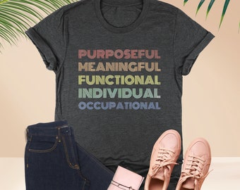 May include: A dark heather grey t-shirt with the words "PURPOSEFUL", "MEANINGFUL", "FUNCTIONAL", "INDIVIDUAL", and "OCCUPATIONAL" printed in rainbow colors.