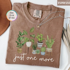 May include: Espresso-colored t-shirt featuring a graphic of various potted plants. The phrase "just one more" is written in white script across the bottom. A silver necklace with a small pendant is draped on the shirt.
