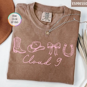 May include: Espresso-colored t-shirt with a pink graphic of a cowboy boot, hat, bow, and horseshoe. The words "Cloud 9" are written in pink script below the graphic. A silver necklace with a pendant is visible.