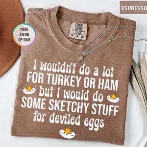 May include: Espresso-colored t-shirt with the text "I wouldn't do a lot FOR TURKEY OR HAM but I would do SOME SKETCHY STUFF for deviled eggs." The shirt has a crew neck and short sleeves. A silver necklace is draped across the shirt.