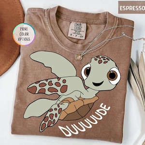 May include: Espresso-colored t-shirt featuring a cartoon turtle with large brown eyes and the word "DUUUUDE" in white. The shirt has a crew neck and short sleeves. A silver necklace with a small pendant is also visible.
