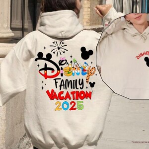 K&ouml;nnte beinhalten: Ein wei&szlig;er Hoodie mit einer schwarzen Mickey-Maus-Silhouette und dem Text "Disney 2025" auf der Vorderseite. Die R&uuml;ckseite des Hoodies zeigt eine bunte Grafik mit dem Text "Disney Family Vacation 2025".