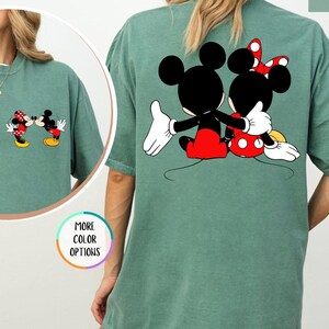 May include: Light green t-shirt with Mickey and Minnie Mouse graphics. A small image of the characters is on the front, and a larger image of them embracing is on the back. The shirt has a relaxed, oversized fit.