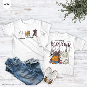 May include: Two white t-shirts with cat graphics and text. One shirt says "everybody wants to be a cat" with cartoon cats. The other says "Bonjour" with a graphic of cats and the Eiffel Tower. Blue jeans and white sneakers are also in the image.