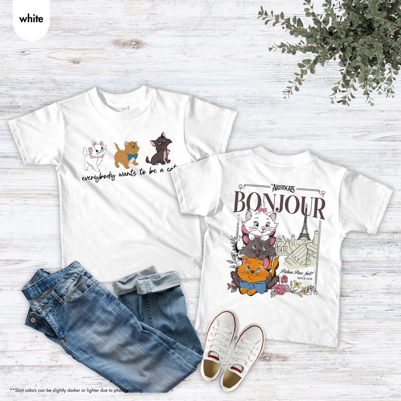 May include: Two white t-shirts with cat graphics and text. One shirt says "everybody wants to be a cat" with cartoon cats. The other says "Bonjour" with a graphic of cats and the Eiffel Tower. Blue jeans and white sneakers are also in the image.