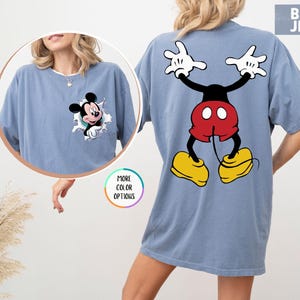 May include: Blue jean coloured t-shirt featuring Mickey Mouse. The front of the shirt has a graphic of Mickey Mouse bursting through a hole. The back of the shirt has a large graphic of Mickey Mouse with his arms up and his legs spread.