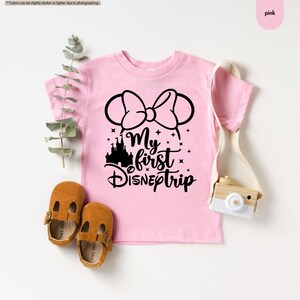 Pu&ograve; includere: Maglietta rosa a maniche corte con una grafica nera di orecchie di Topolino con un fiocco, un castello e il testo "My first Disney trip".