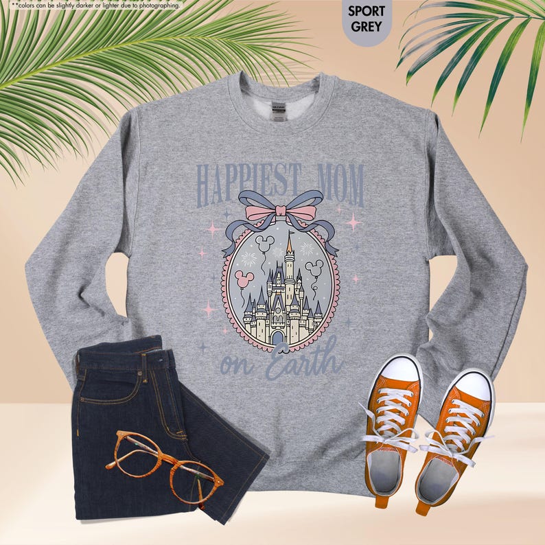 May include: A gray sweatshirt with the text "Happiest Mom on Earth" and a castle graphic. The sweatshirt is paired with dark blue jeans, orange-framed glasses, and orange sneakers.