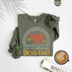 Puede incluir: Una sudadera verde con un gr&aacute;fico de estilo vintage de una figura musculosa con el texto "Just an Ordinary Demi-Dad".